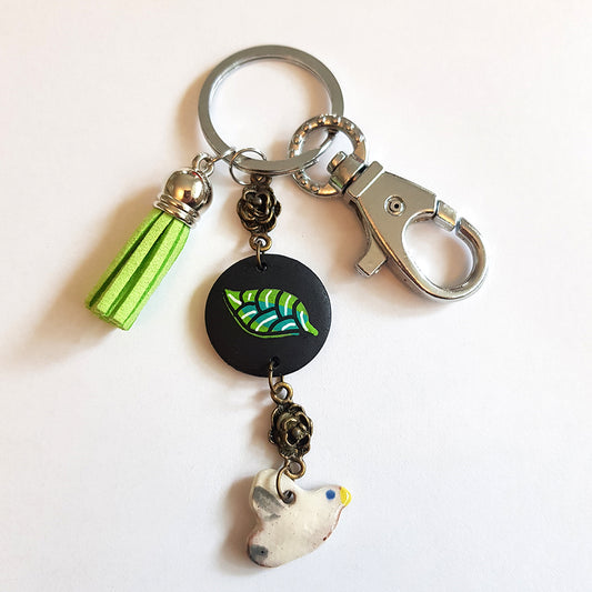 A stylish CocoFlower Handmade Bird keychain with a green tassel, a metallic bead, a black charm with a green leaf design, and a quirky white ceramic charm shaped like a bird with blue and yellow details, all attached to a silver key ring with a lobster clasp.