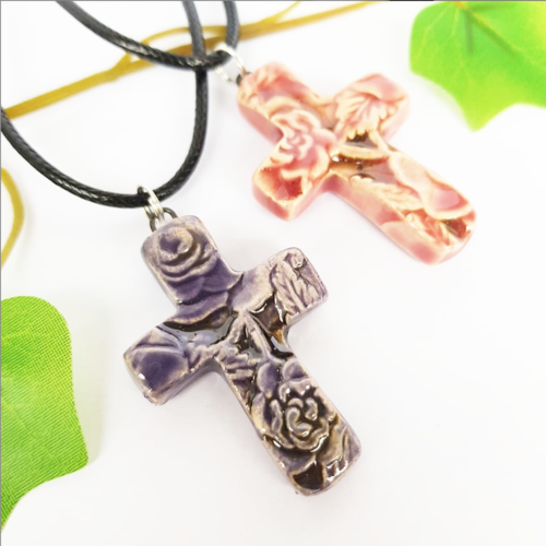 Two cocoflower ceramic cross necklaces featuring floral designs suspended on a black cord, with a green leafy backdrop suggesting peace and spirituality.