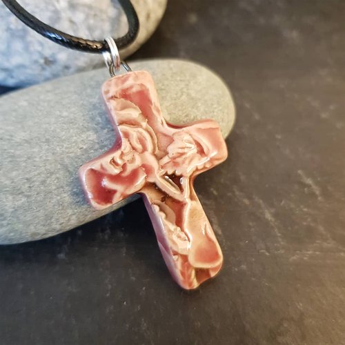 An artisan pink ceramic cross pendant with an embossed floral design resting on a gray wooden surface