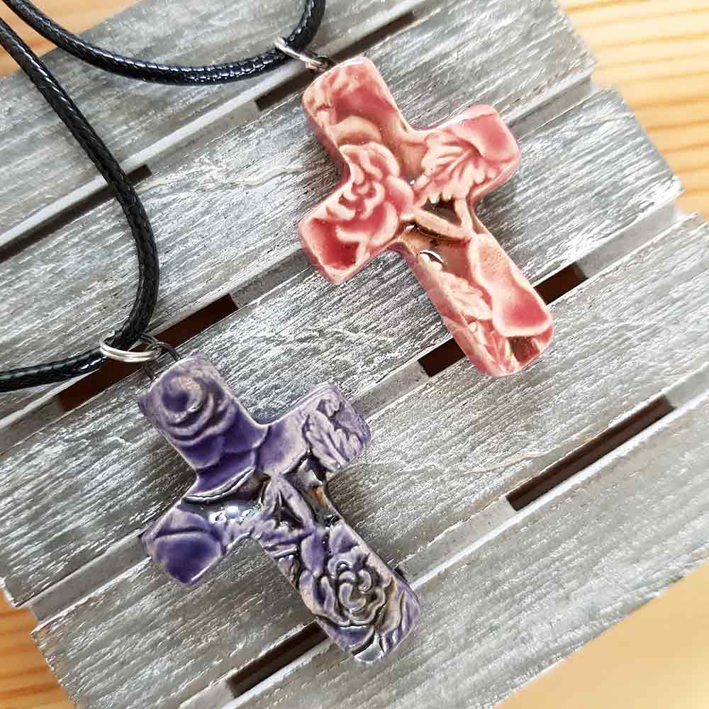 Two CocoFlower handmade ceramic necklace cross pendants, one in shades of pink and the other in purple, displayed on a textured gray surface.